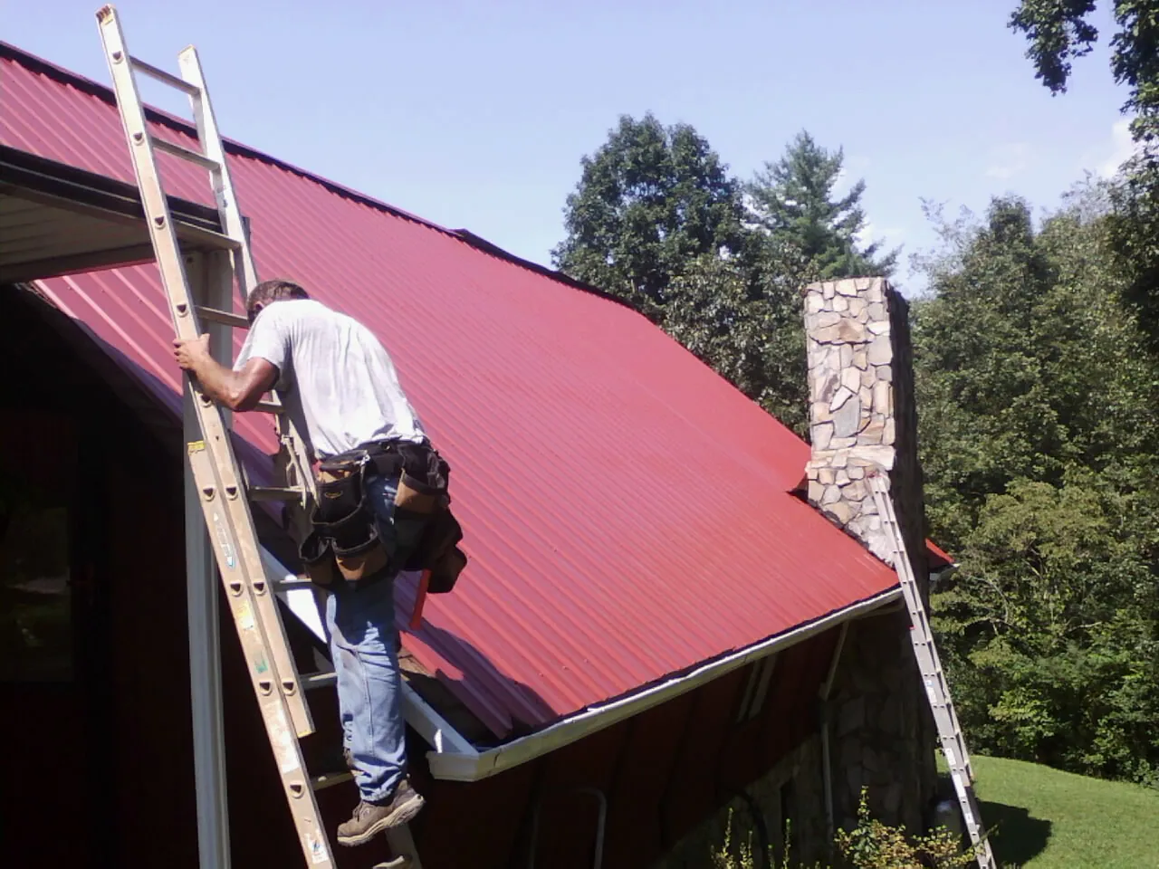Roofer on a ladder beside a red metal roof and stone chimney — Soffit & Fascia Repair by Roofing Brothers in East Stone Gap